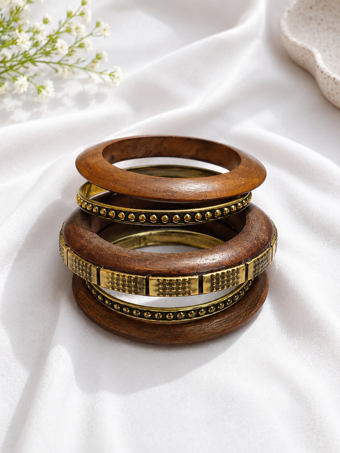 Stack of four wood and brass bangles resting on white draped fabric with baby's breath flowers in background