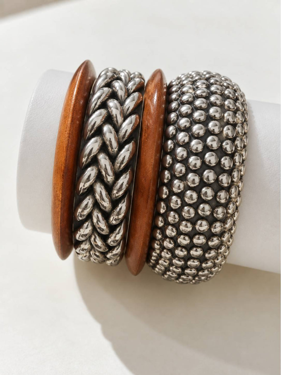 Three bangles stacked on a white cylindrical display — two oxidised silver metal bangles and one smooth wooden bangle