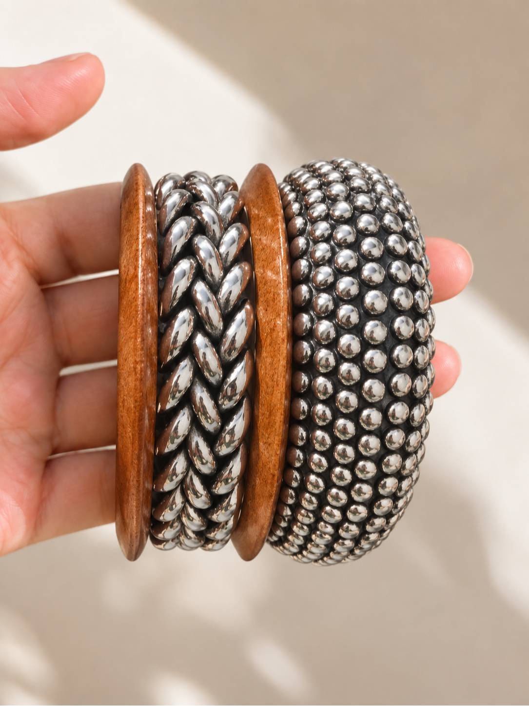 Hand holding three stacked bangles — wood bangle flanked by braided and ball-stud oxidised silver bangles, in natural sunlight