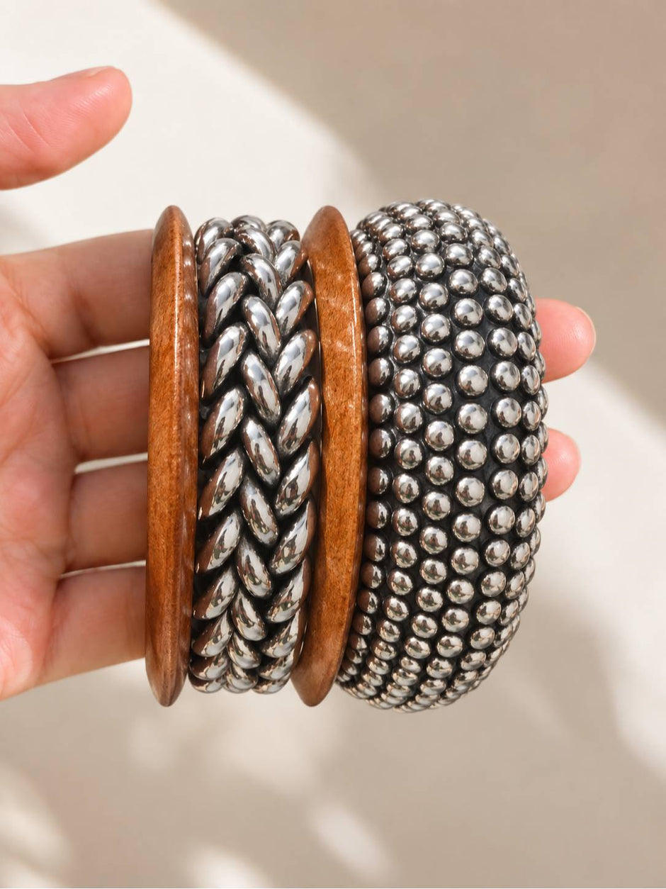 Hand holding three stacked bangles — wood bangle flanked by braided and ball-stud oxidised silver bangles, in natural sunlight