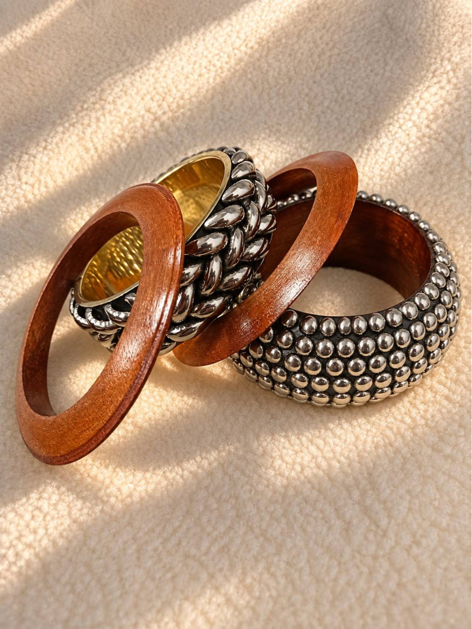 Three bangles laid flat on a cream textured surface — two wooden bangles and one braided silver bangle with gold interior, in dappled light