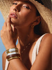 Model wearing gold-toned bangle stack with a wide-brim straw hat and ivory top in bright sunlight
