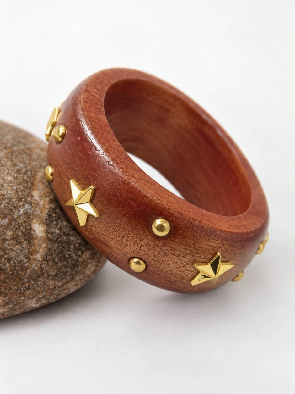 Wide wooden bangle with gold star and dome studs resting against a rounded grey stone on a white surface