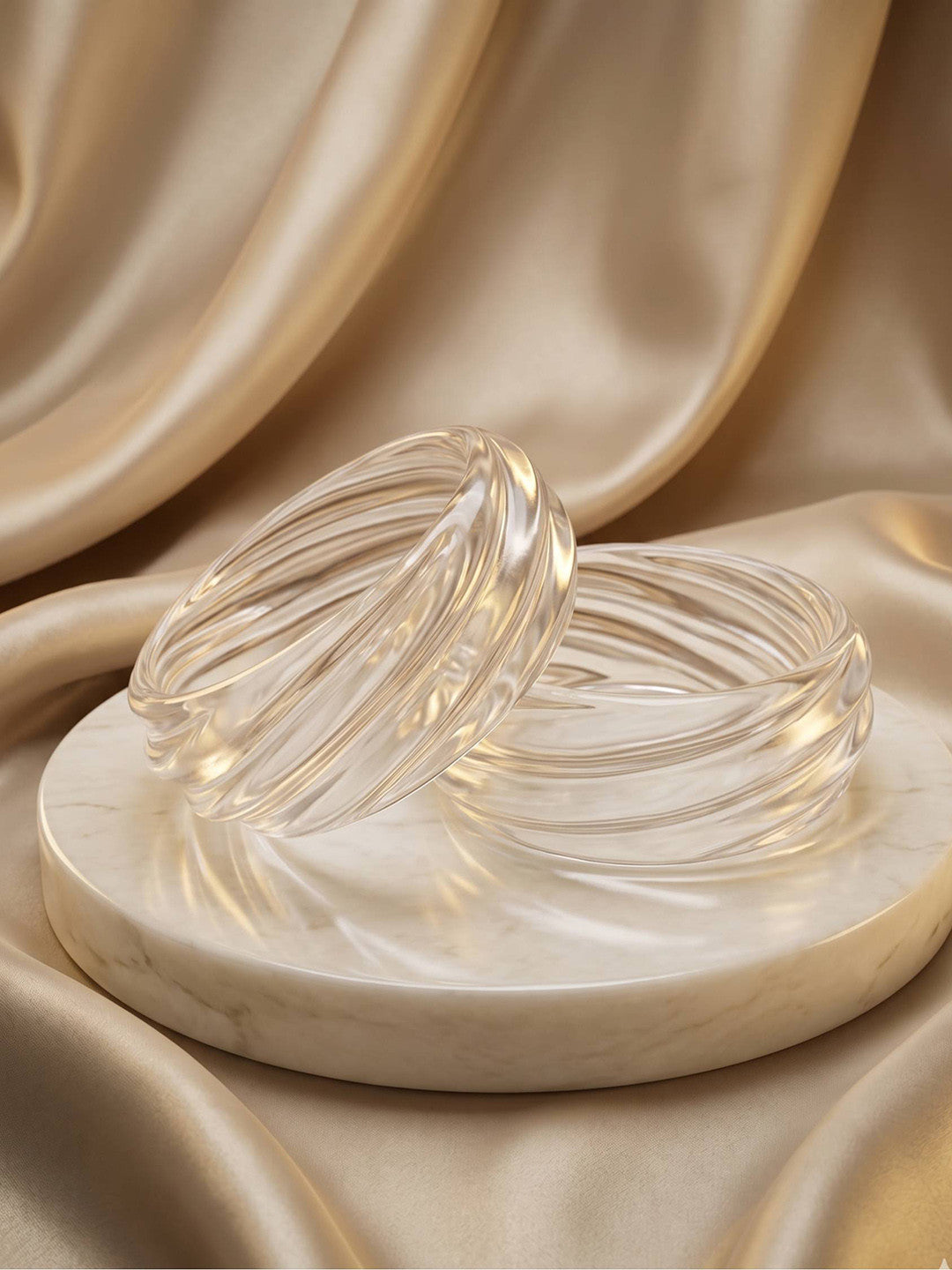 Two clear textured resin bangles with swirled ribbing displayed on a marble disc against champagne satin