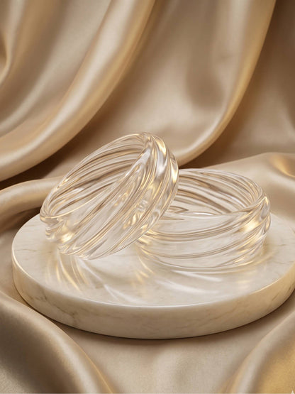 Two clear textured resin bangles with swirled ribbing displayed on a marble disc against champagne satin