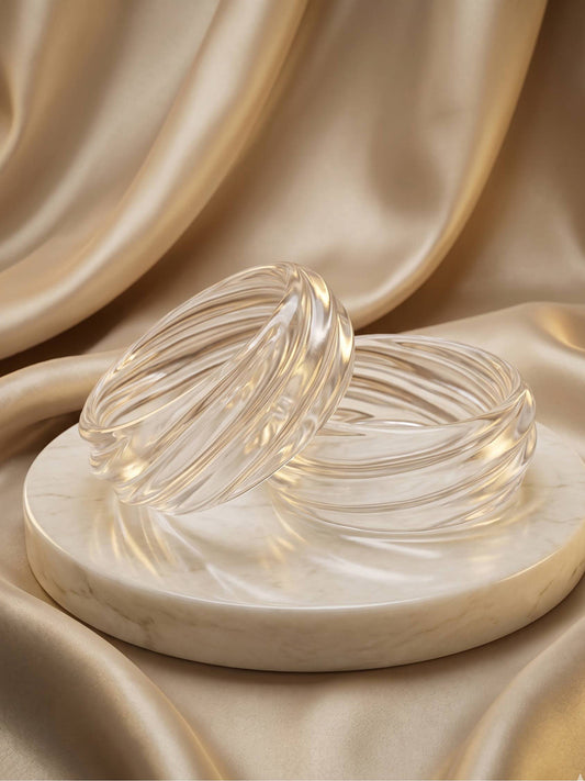Two clear textured resin bangles with swirled ribbing displayed on a marble disc against champagne satin