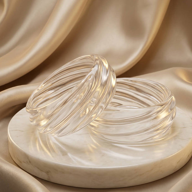 Two clear textured resin bangles with swirled ribbing displayed on a marble disc against champagne satin