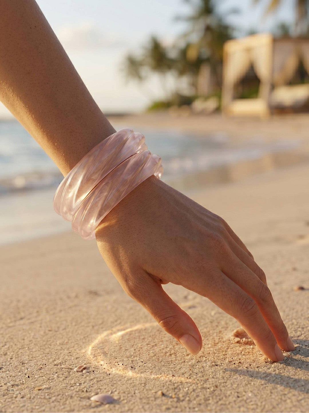 Clear resin bangles stacked on a wrist with fingers tracing a heart in golden beach sand at sunset