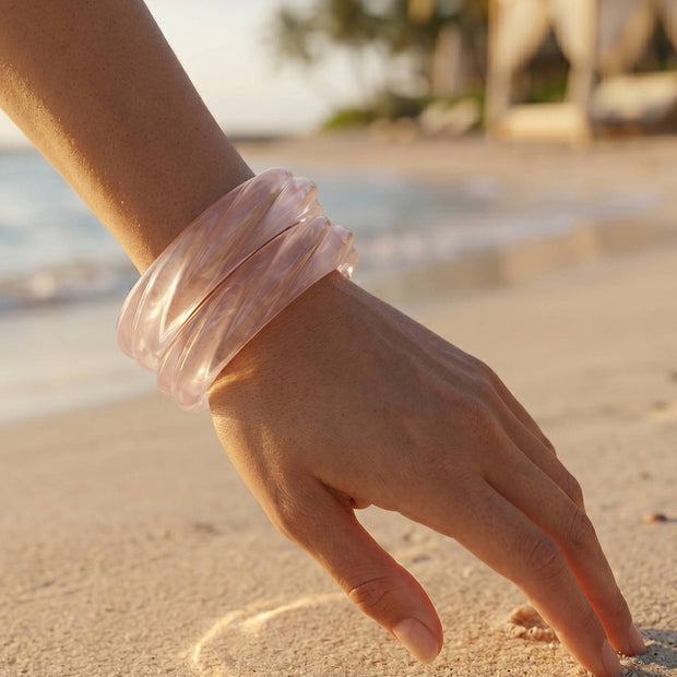 Clear resin bangles stacked on a wrist with fingers tracing a heart in golden beach sand at sunset