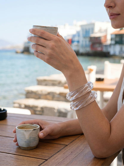 Clear ribbed resin bangles stacked on a wrist as a woman holds a ceramic cup at a waterfront café in Mykonos
