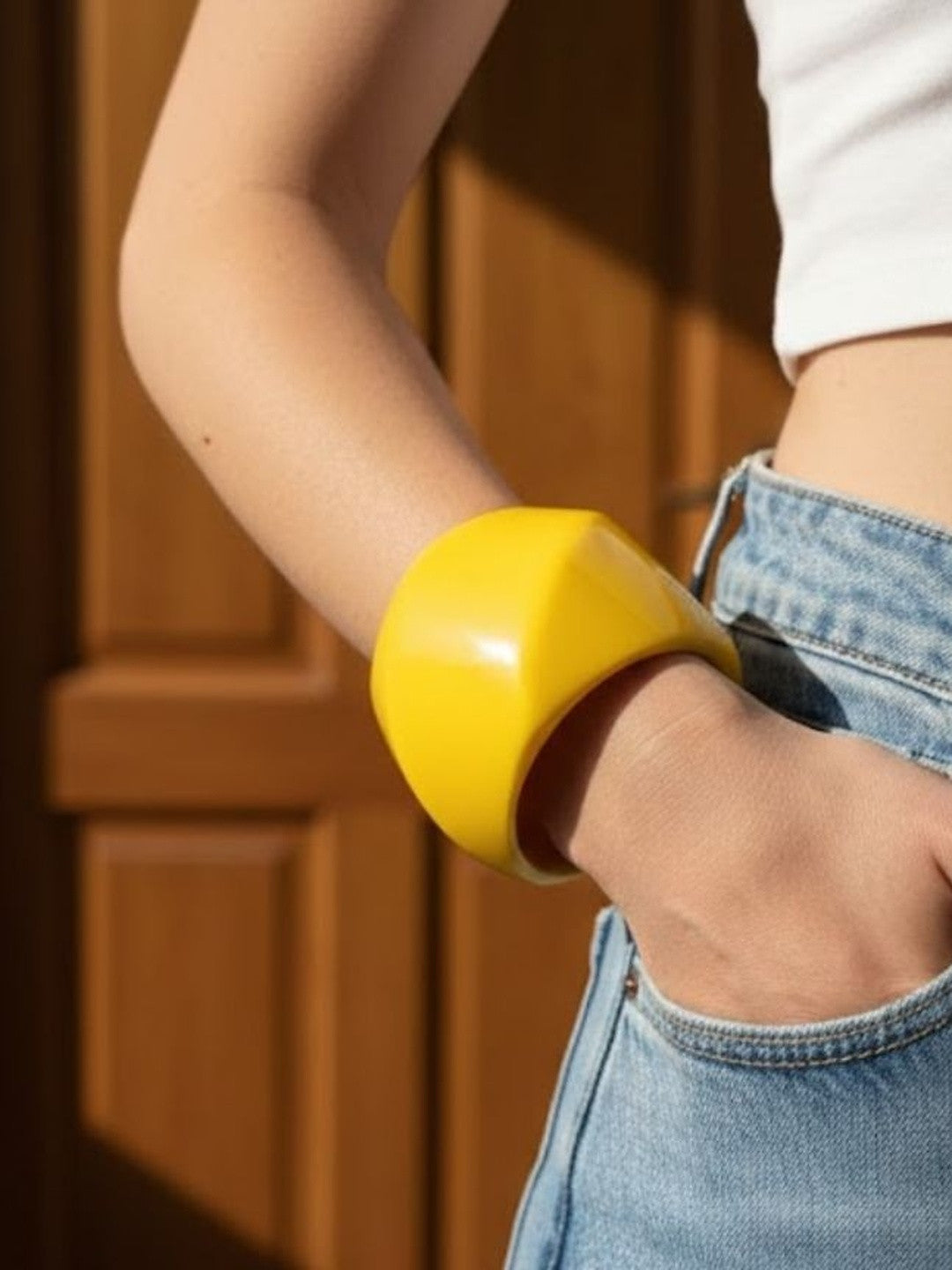 Chunky yellow resin cuff bangle worn on wrist of woman in white crop top and blue denim jeans