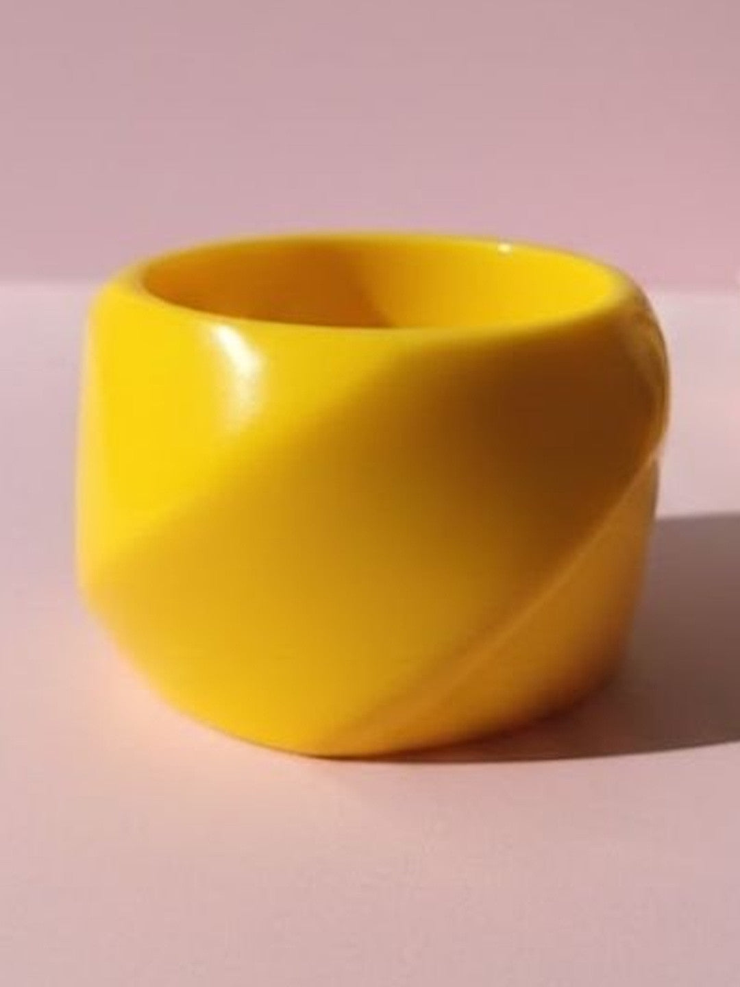 Glossy yellow resin cuff bangle with twisted sculptural form photographed on a soft pink surface