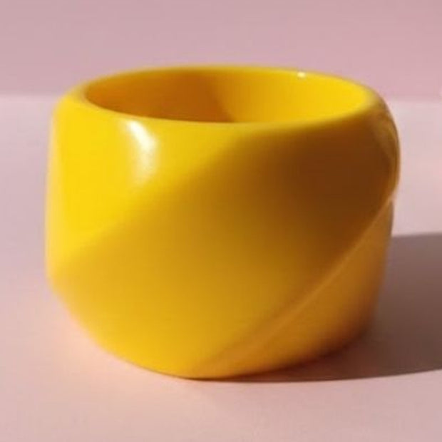 Glossy yellow resin cuff bangle with twisted sculptural form photographed on a soft pink surface
