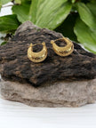 Pair of gold-plated hammered chunky hoop earrings resting on a dark rock with green leaves in background