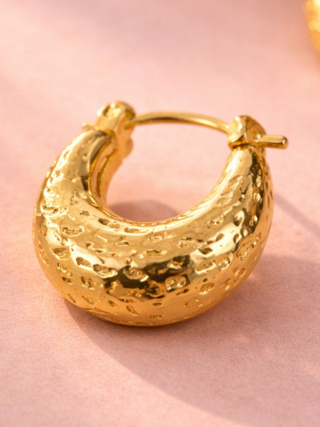 Close-up of a single gold-plated hammered chunky hoop earring showing snap closure and dimpled texture on pink background