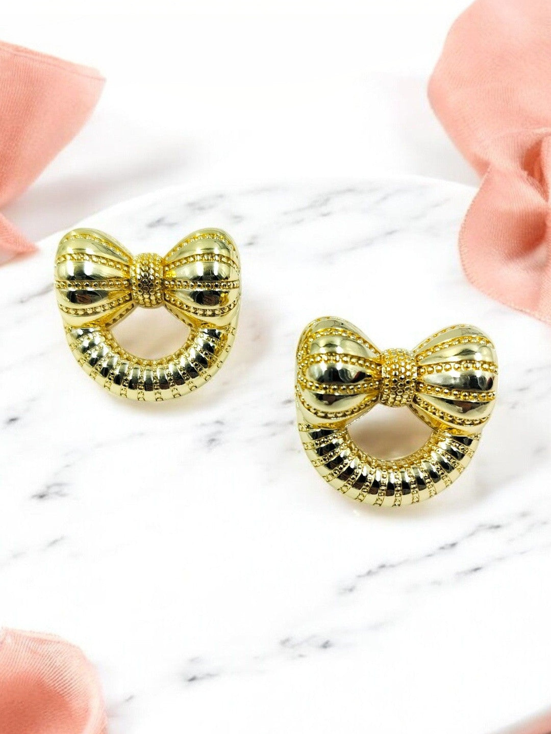 Pair of gold-toned bow stud earrings with ribbed texture on white marble surface with pink ribbon