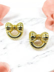 Pair of gold-toned bow stud earrings with ribbed texture on white marble surface with pink ribbon