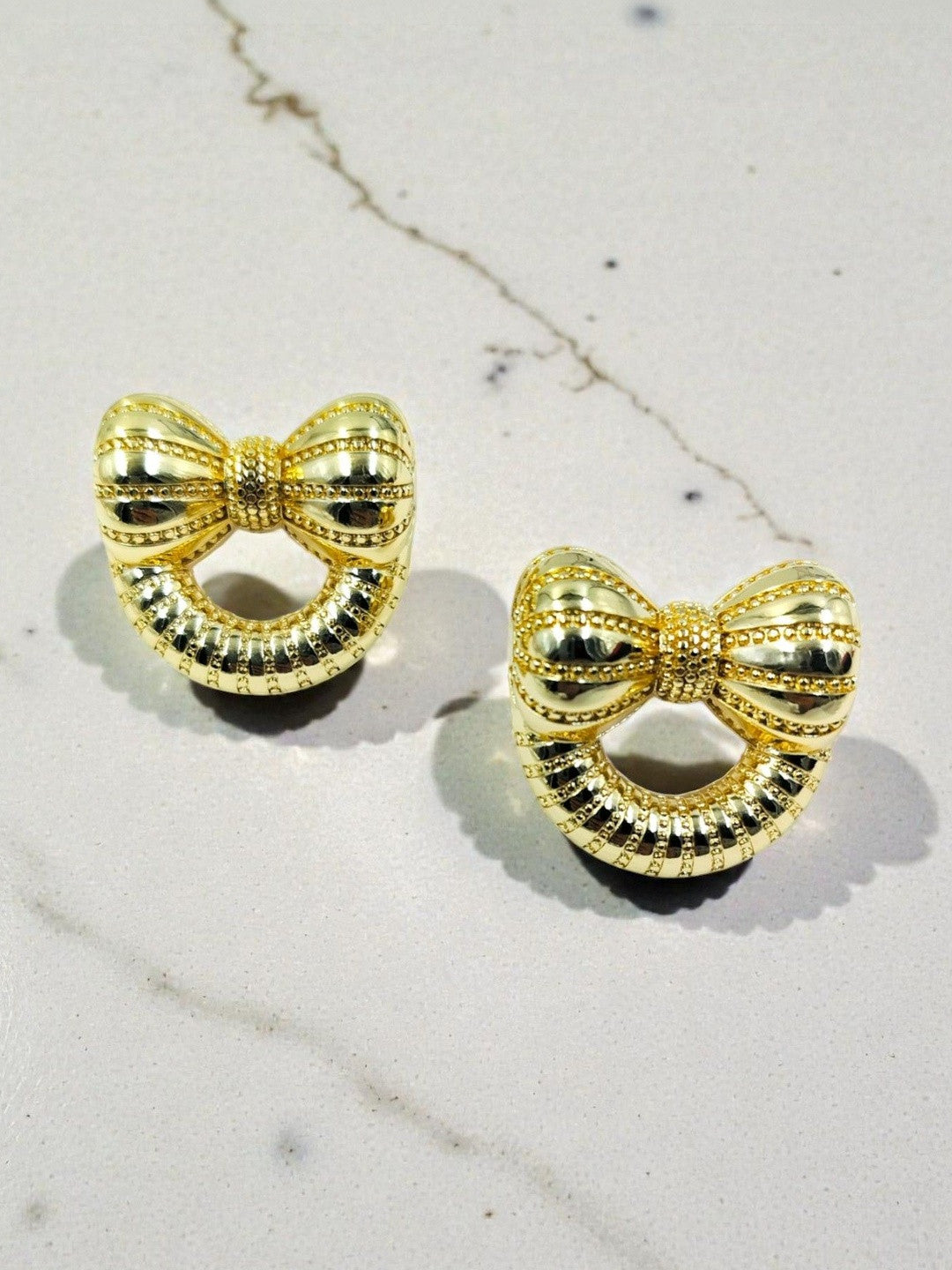 Two gold bow earrings with beaded border and open centre placed on grey marble background