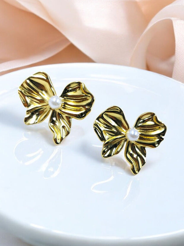 Pair of gold-plated bow stud earrings with pearl centre on white plate, pink ribbon background