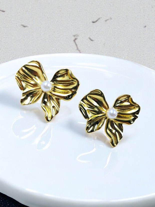 Gold bow stud earrings with faux pearl accent displayed on white ceramic plate, neutral background