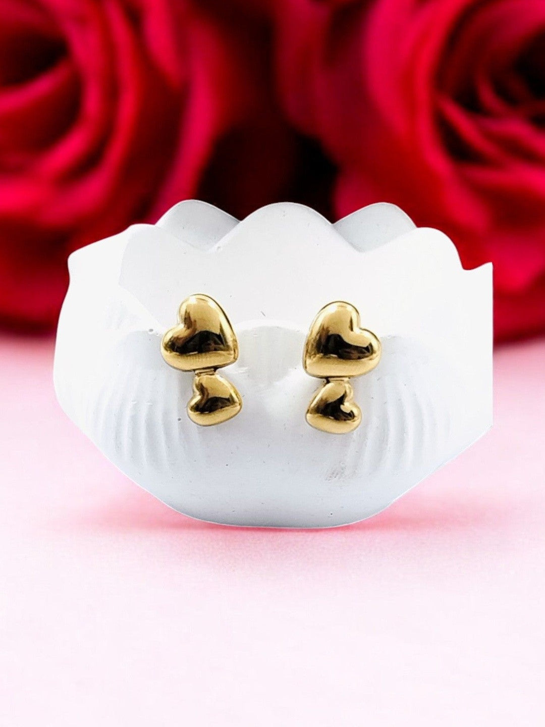 Pair of gold-plated double heart stud earrings displayed on white shell dish with red roses in background