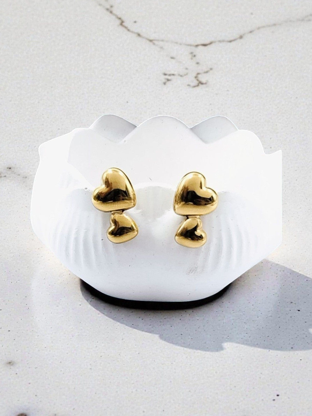 Gold-tone double heart stud earrings resting in white scalloped dish on marble surface in natural light