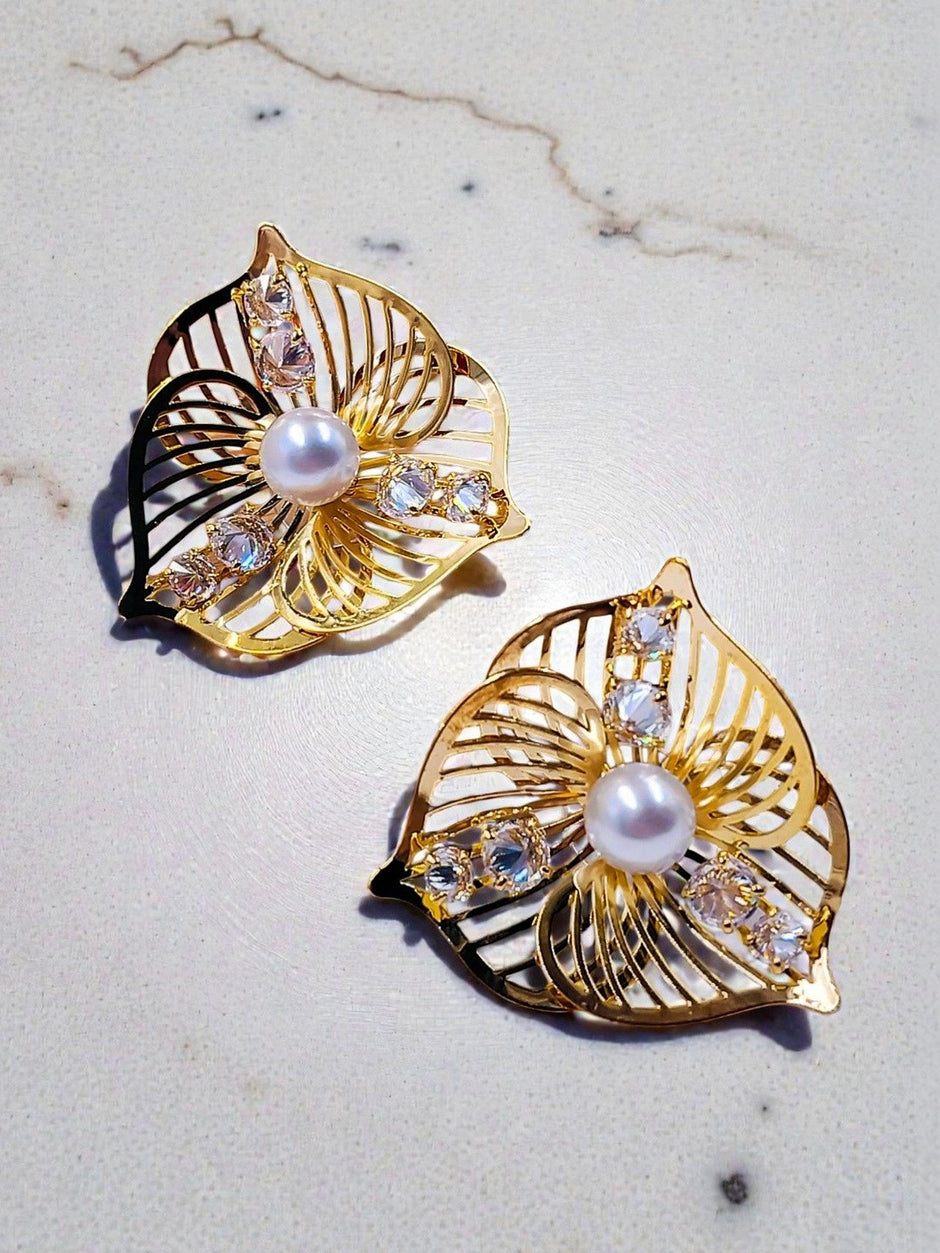 Two gold-plated hollow leaf earrings with faux pearl and clear crystals laid flat on a white marble surface