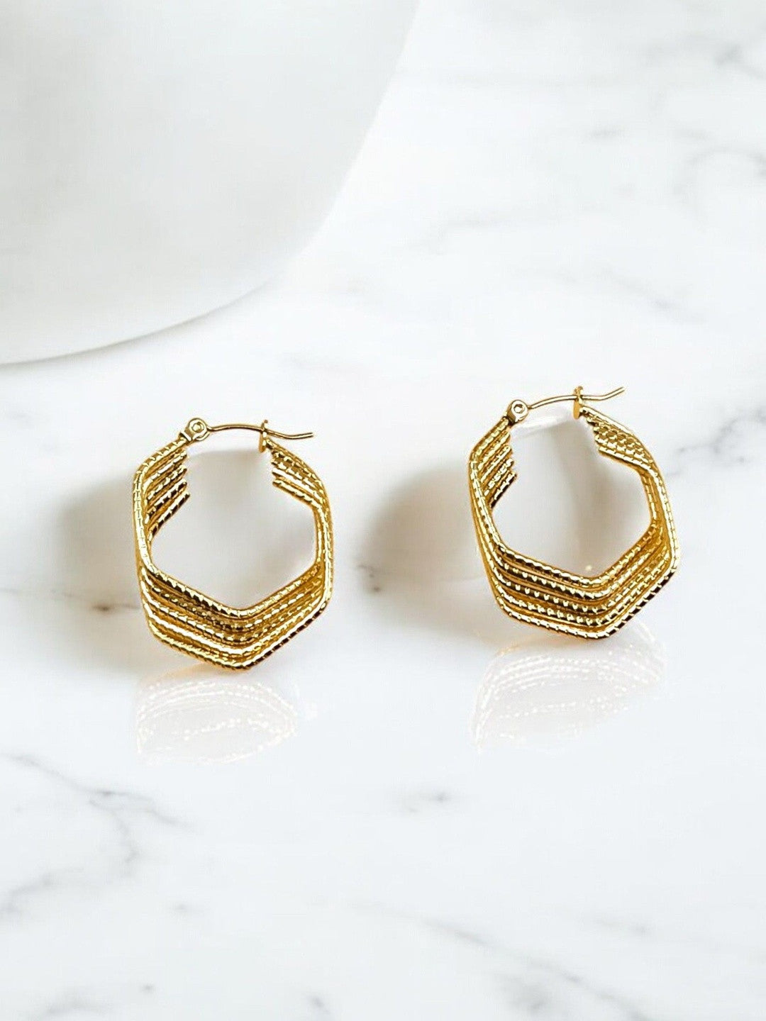 Pair of gold-plated geometric hoop earrings with ridged layered texture on a white marble surface
