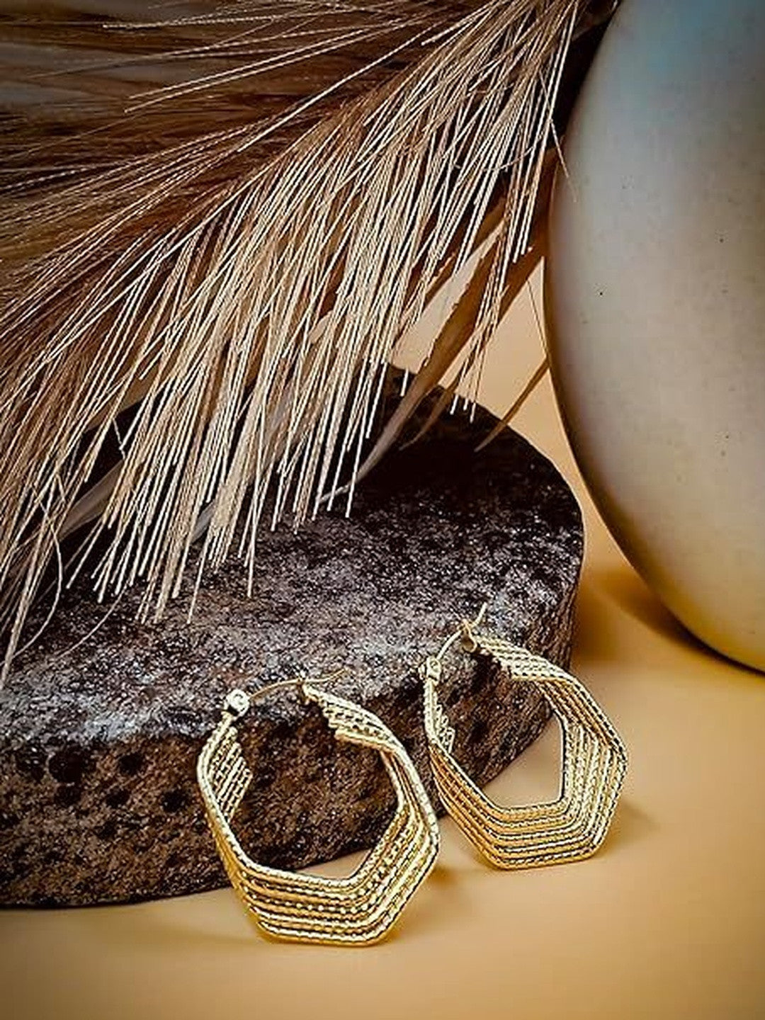 Gold ridged geometric hoop earrings resting on a dark stone with dried grass and ceramic in background