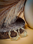 Gold ridged geometric hoop earrings resting on a dark stone with dried grass and ceramic in background