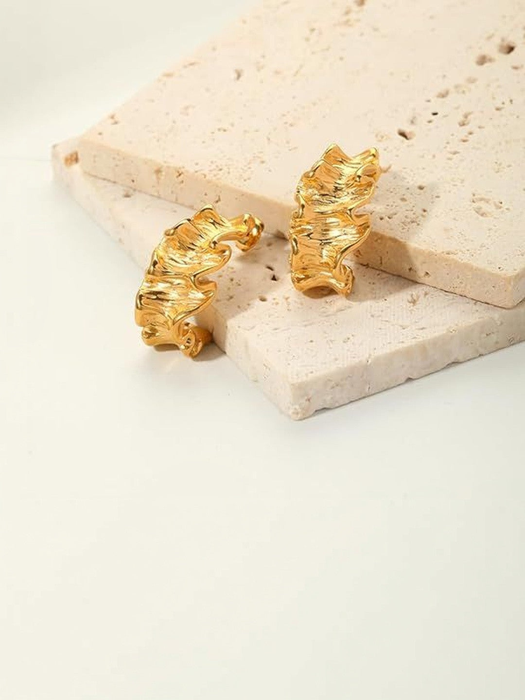 Pair of gold-plated ruffled hoop earrings resting against a beige travertine stone surface