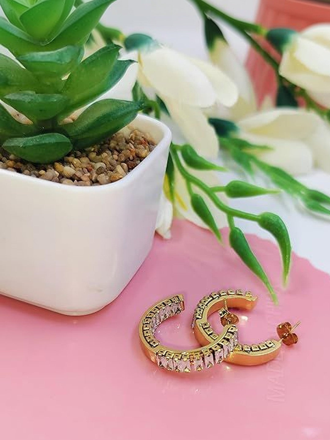 Two gold CZ baguette hoop earrings lying flat on a pink surface beside a succulent plant