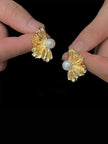 Two hands holding gold floral pearl stud earrings against a black background showing detail