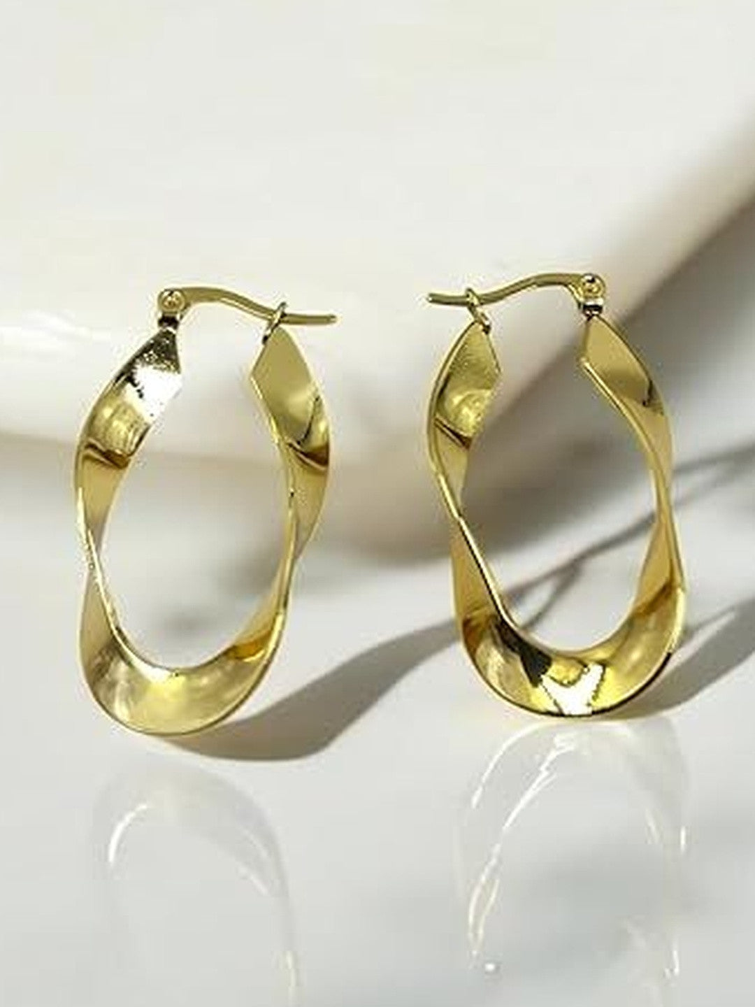 Pair of gold-plated twisted oval hoop earrings resting against a white marble surface