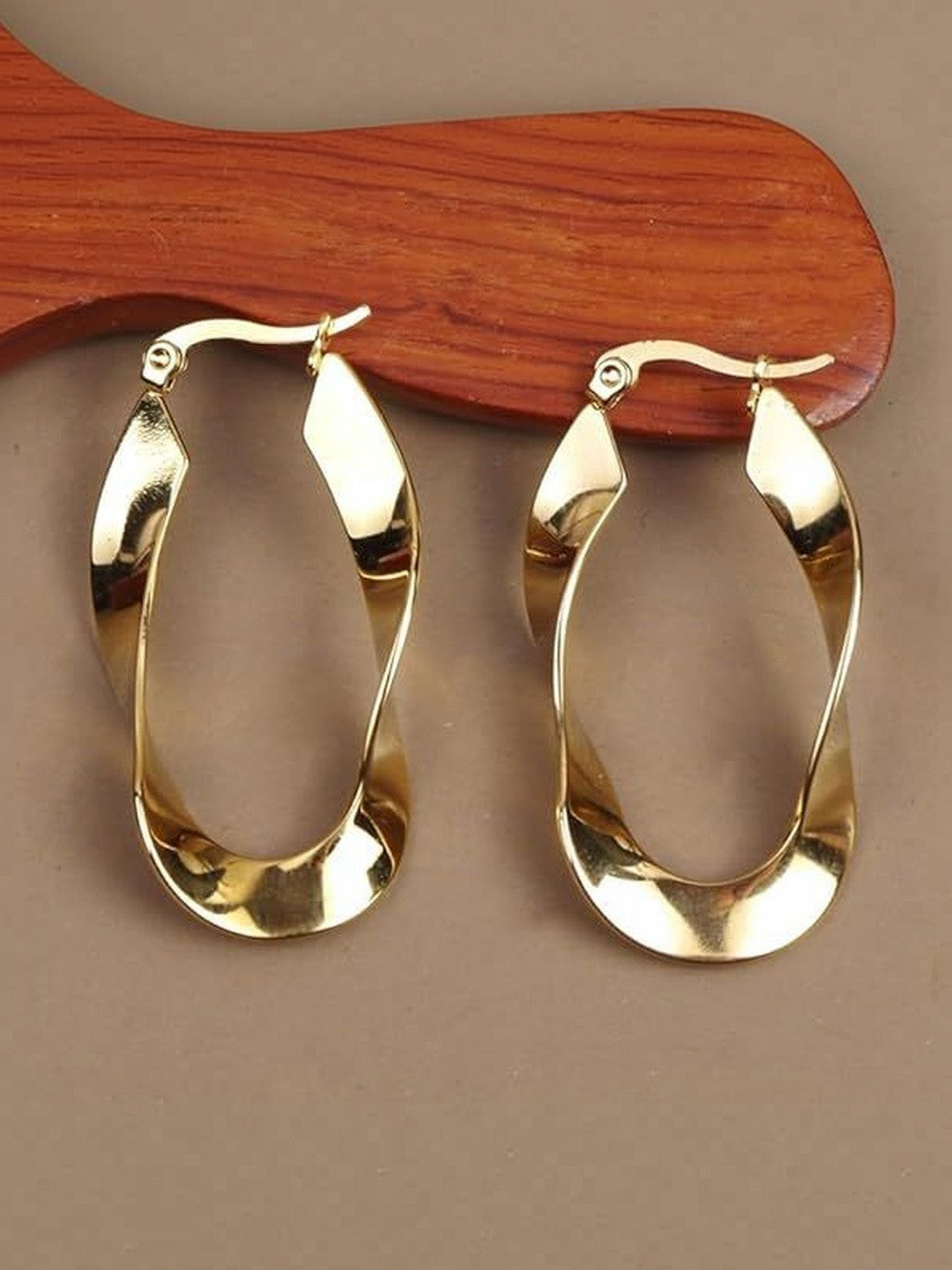 Two gold-tone twisted ribbon hoop earrings displayed on a warm wooden prop