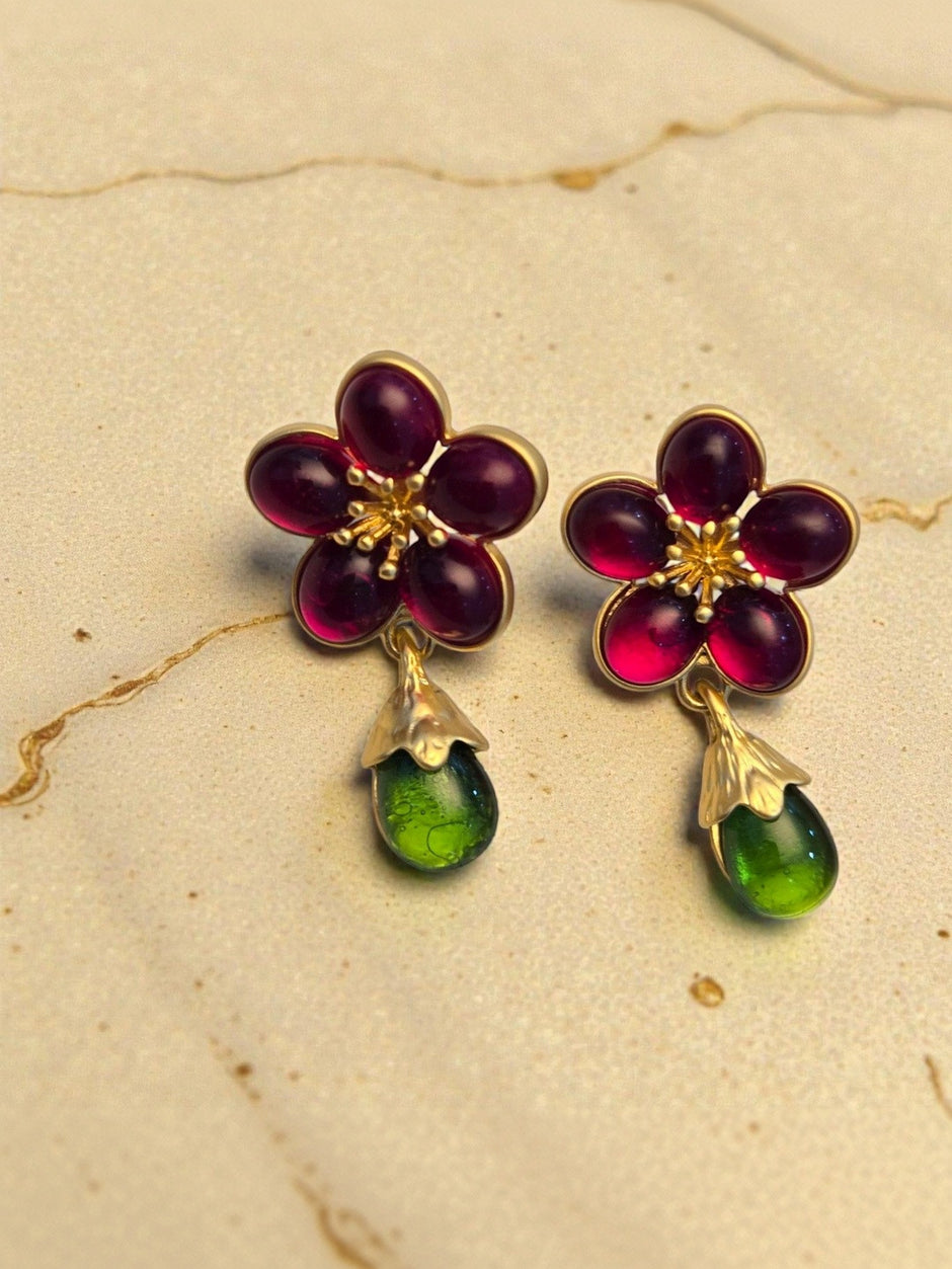 Close-up of ruby-red five-petal glass flower earrings with gold stamen detail and hanging green glass drop on warm beige background