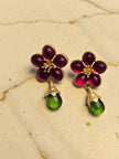 Close-up of ruby-red five-petal glass flower earrings with gold stamen detail and hanging green glass drop on warm beige background