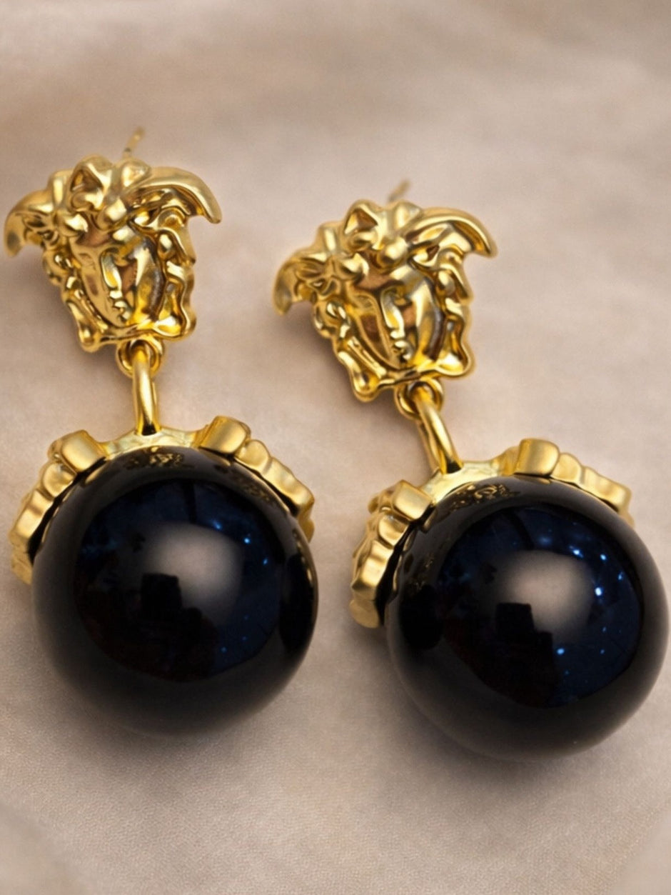 Pair of gold-plated Medusa face stud earrings with large black shimmer ball drops on beige fabric
