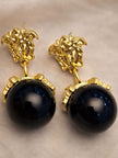 Pair of gold-plated Medusa face stud earrings with large black shimmer ball drops on beige fabric