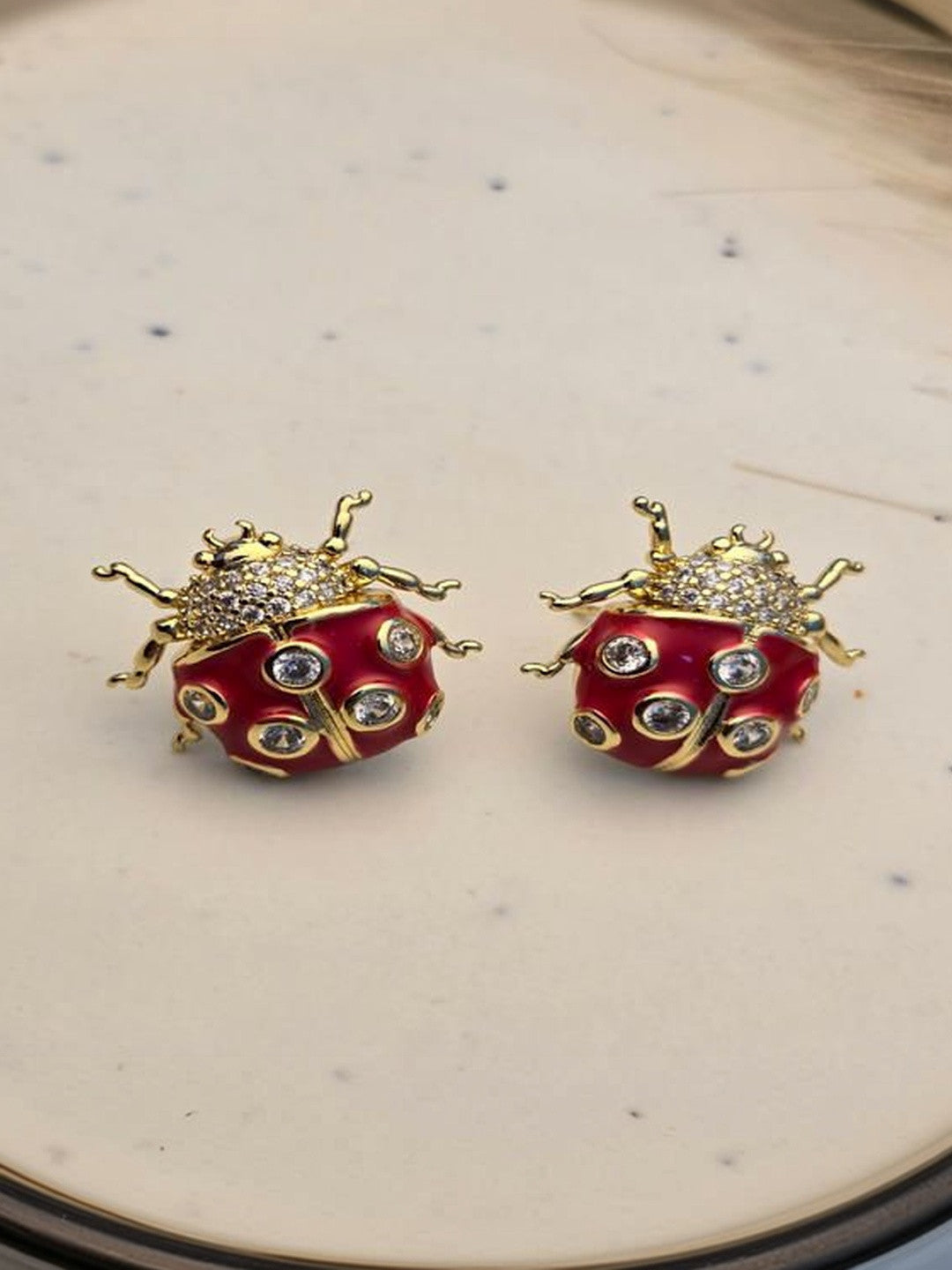 Pair of gold-plated red enamel ladybug stud earrings with cubic zirconia spots on a cream ceramic surface
