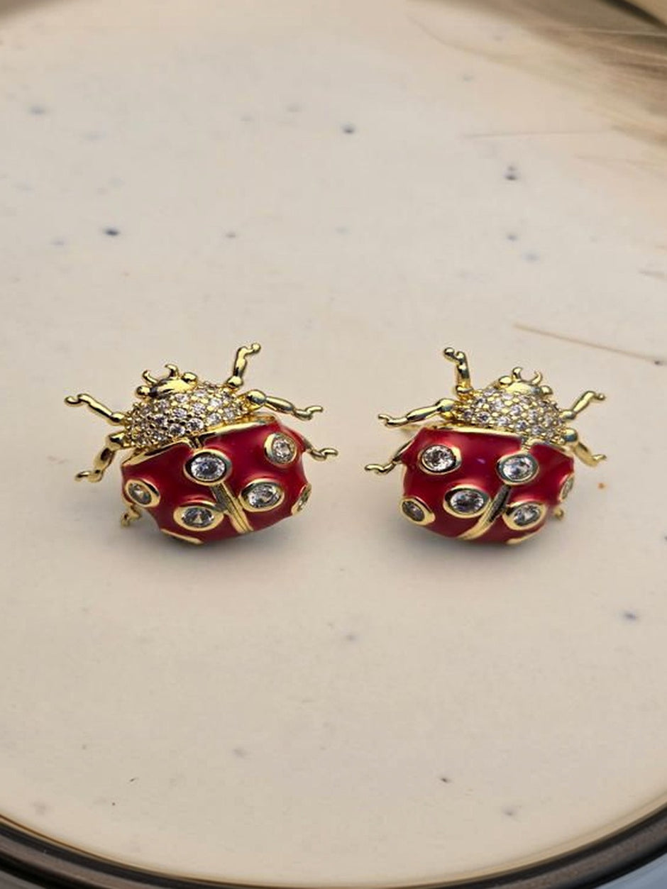 Pair of gold-plated red enamel ladybug stud earrings with cubic zirconia spots on a cream ceramic surface