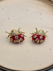 Pair of gold-plated red enamel ladybug stud earrings with cubic zirconia spots on a cream ceramic surface