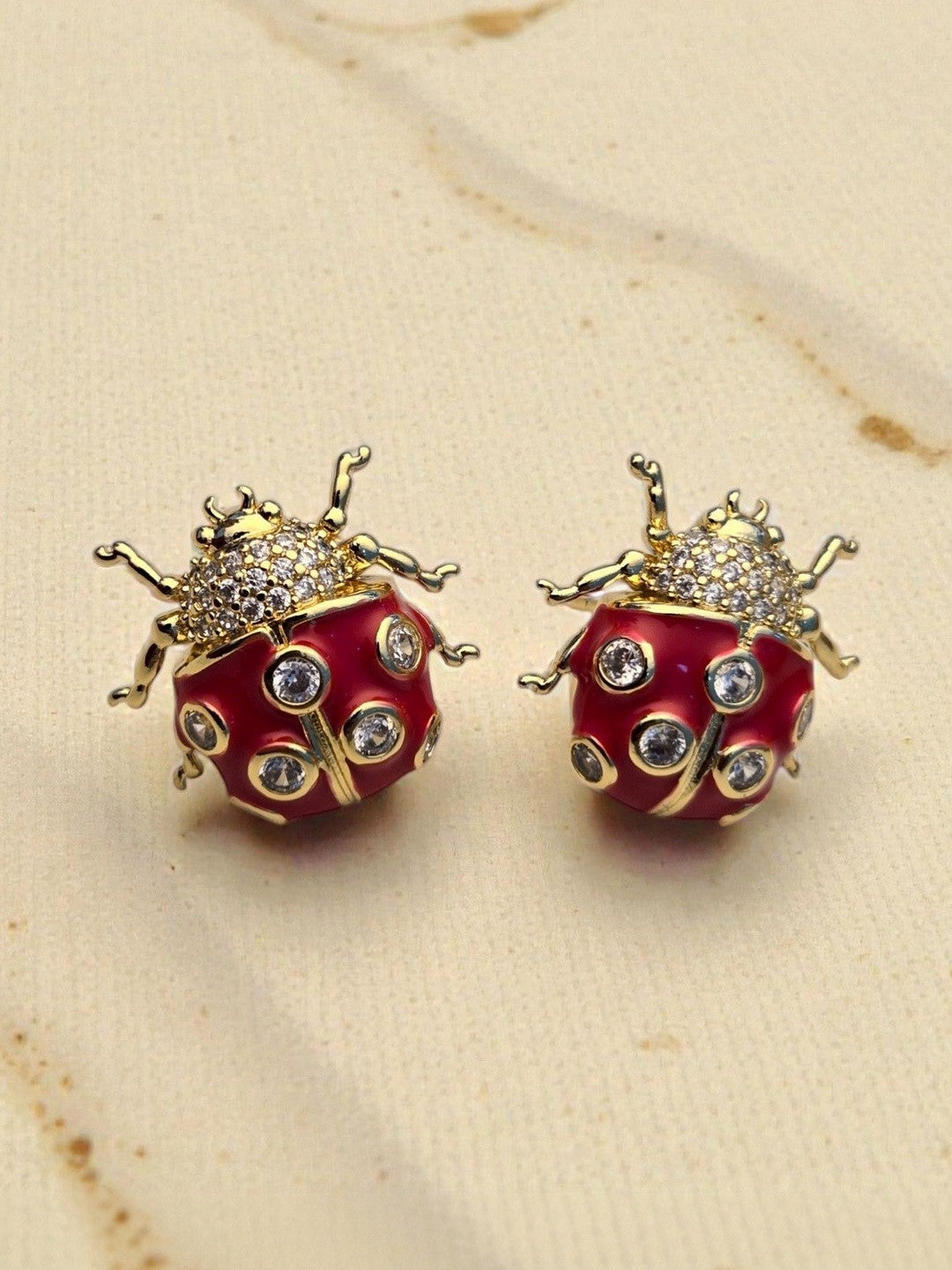 Close-up of two gold-toned red enamel ladybug earrings showing CZ detail and leg texture on a linen surface