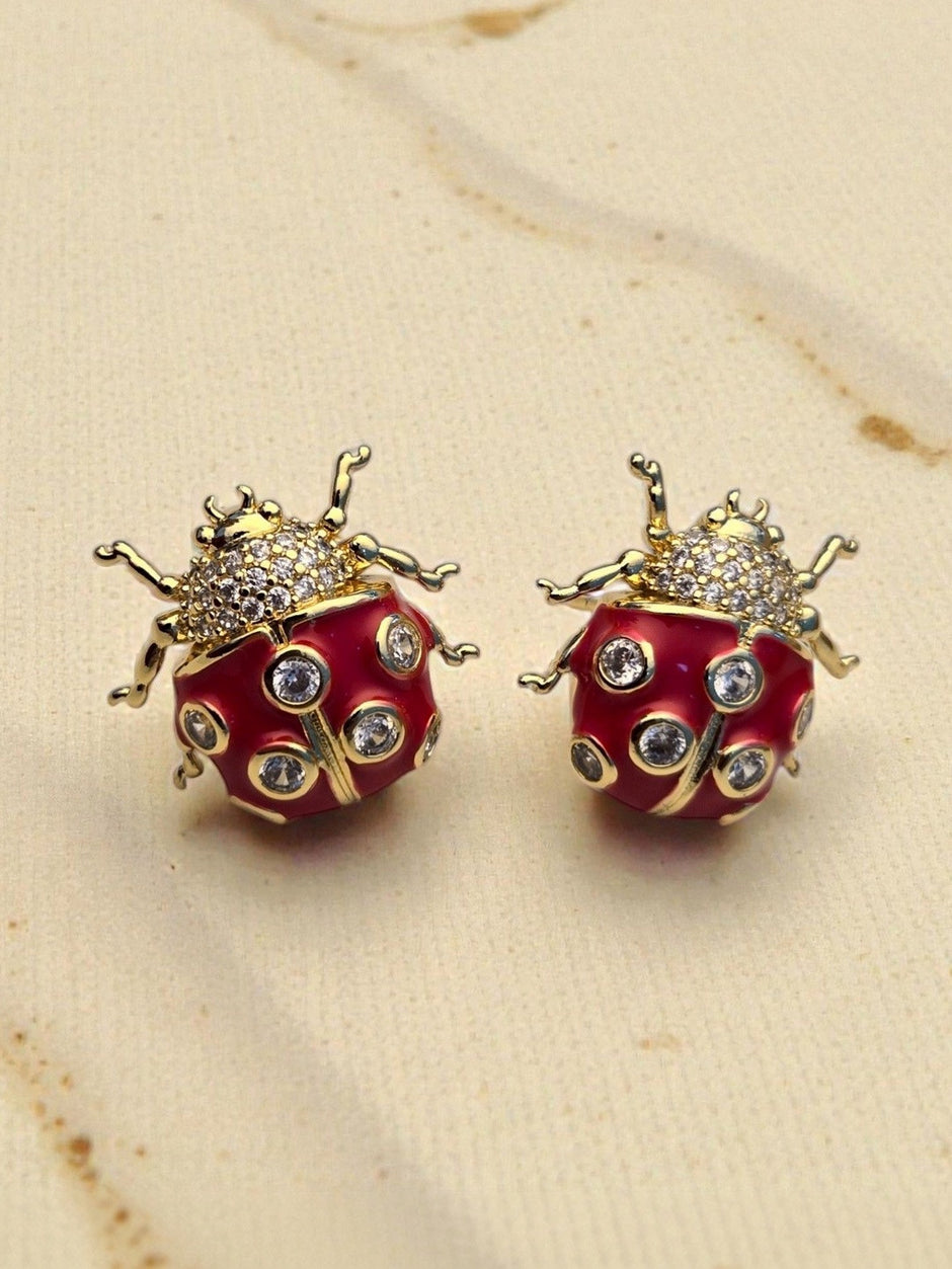 Close-up of two gold-toned red enamel ladybug earrings showing CZ detail and leg texture on a linen surface