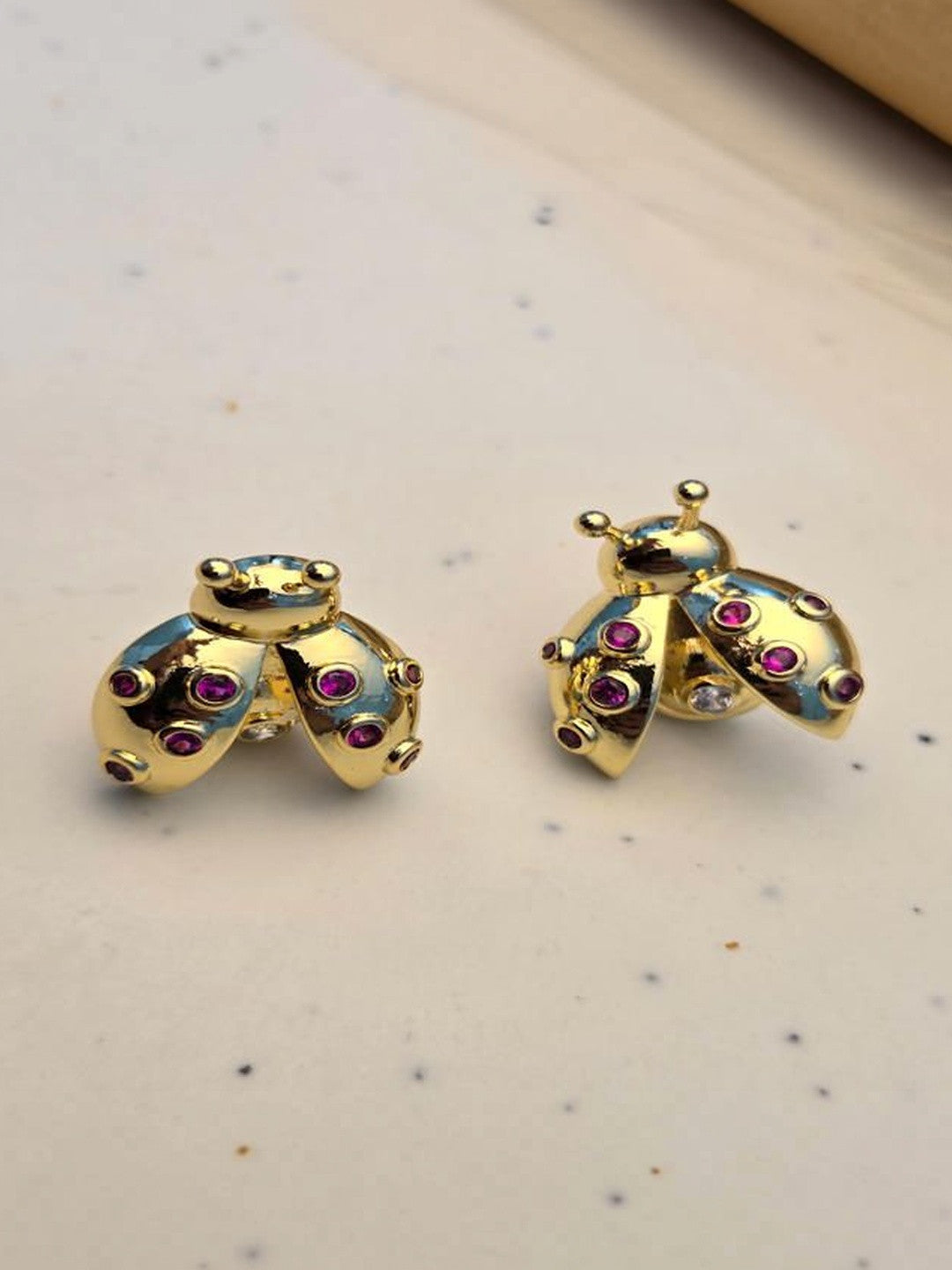 Pair of gold-plated ladybug stud earrings with ruby-red zirconia on a white speckled surface