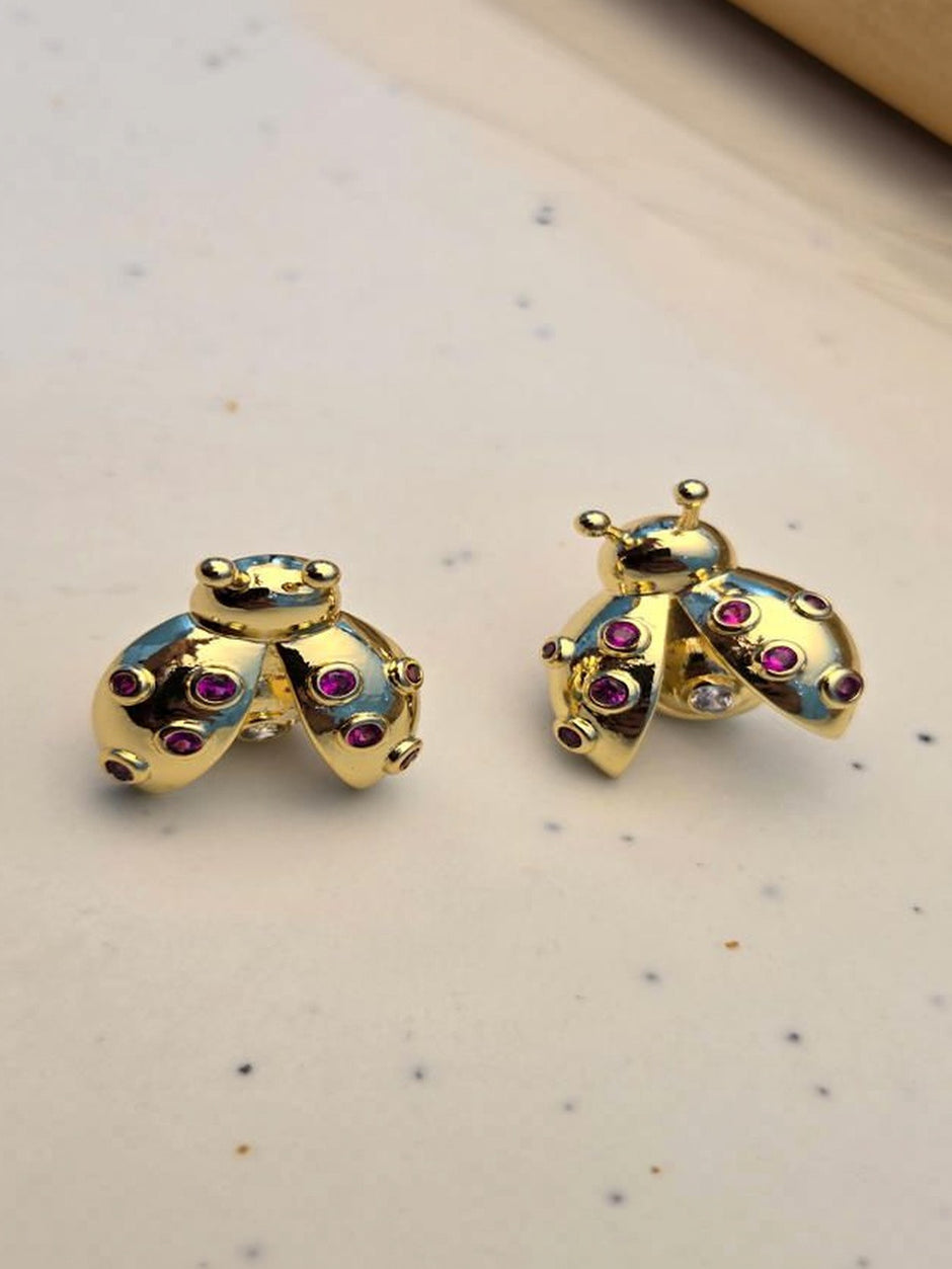 Pair of gold-plated ladybug stud earrings with ruby-red zirconia on a white speckled surface