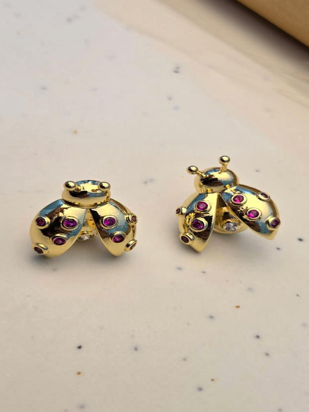 Pair of gold-plated ladybug stud earrings with ruby-red zirconia on a white speckled surface