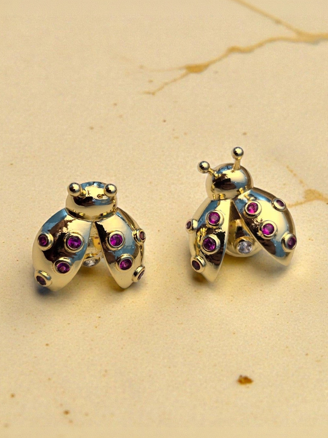 Close-up of gold-plated ladybug stud earrings showing bezel-set red zirconia on a cream marble surface