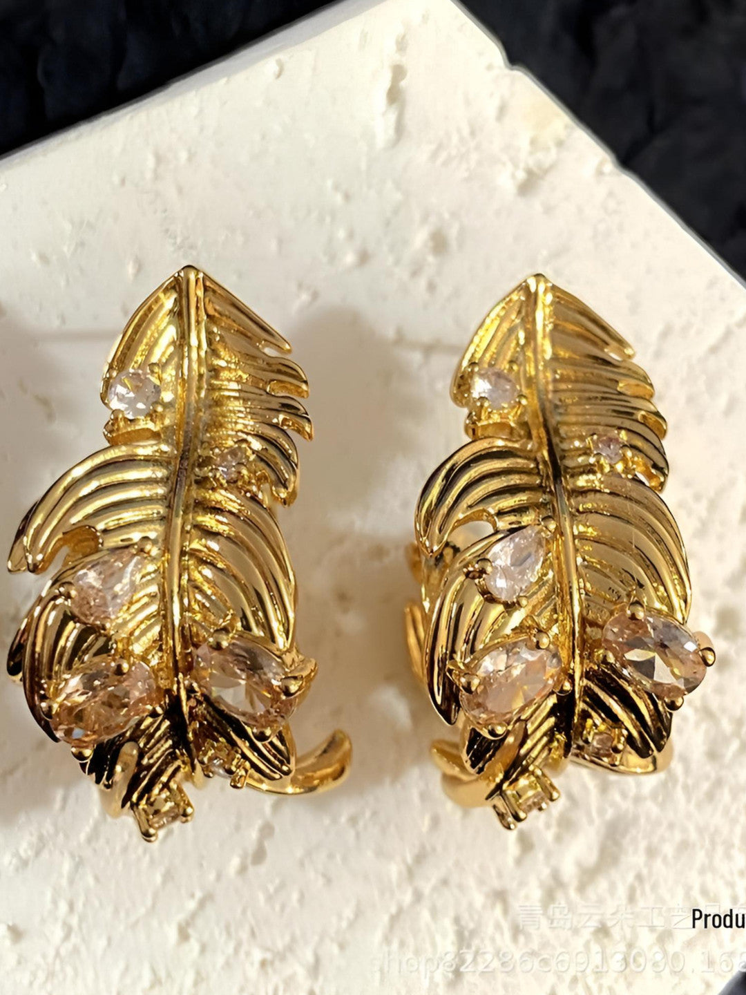 Pair of gold-plated feather stud earrings with champagne cubic zirconia stones on a white display surface