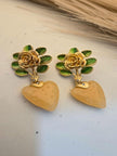 Pair of gold-plated rose and green enamel leaf earrings with peach resin heart drops, placed on a white surface near dried grass